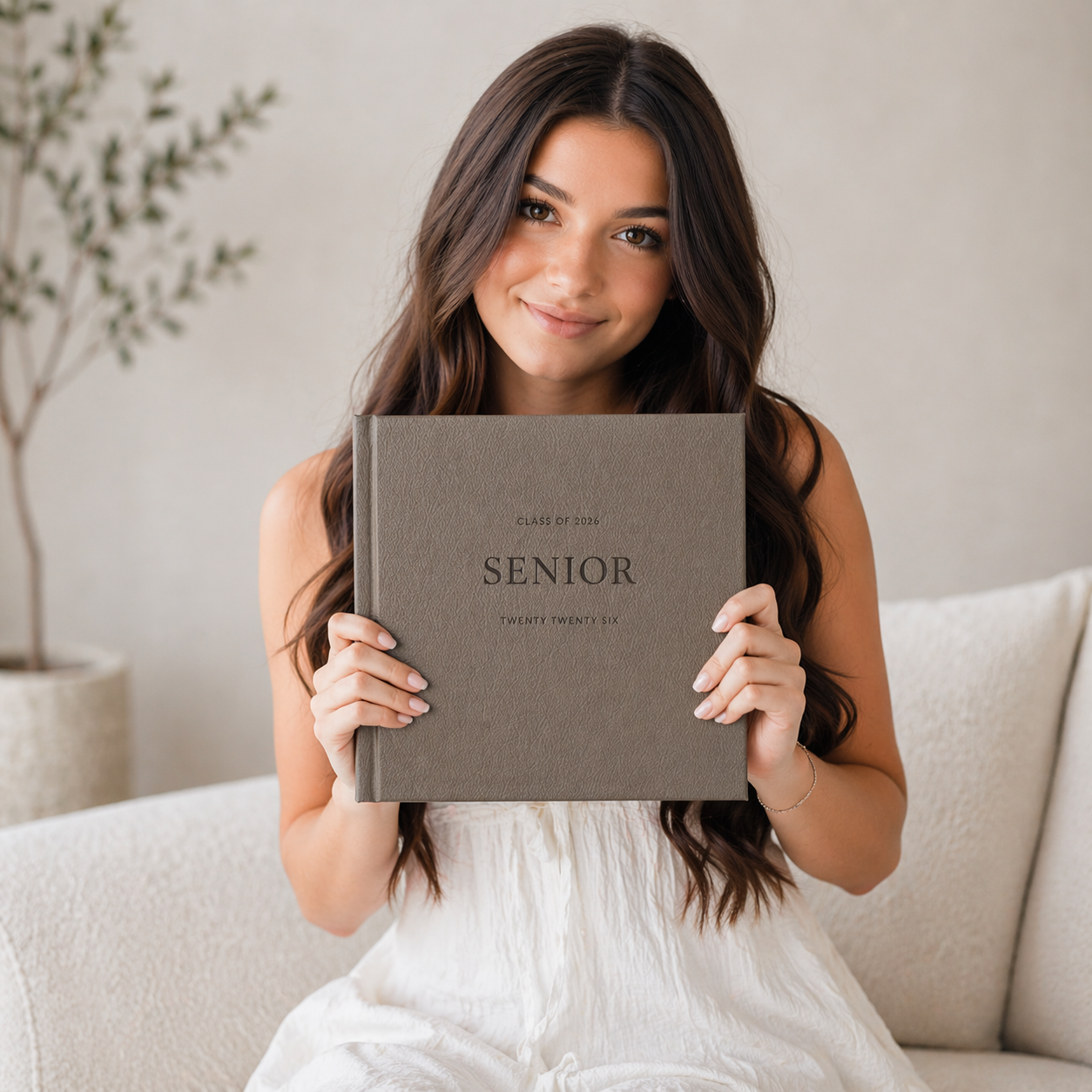 Young woman in white dress holding a taupe linen Senior memory book while sitting on a white couch.