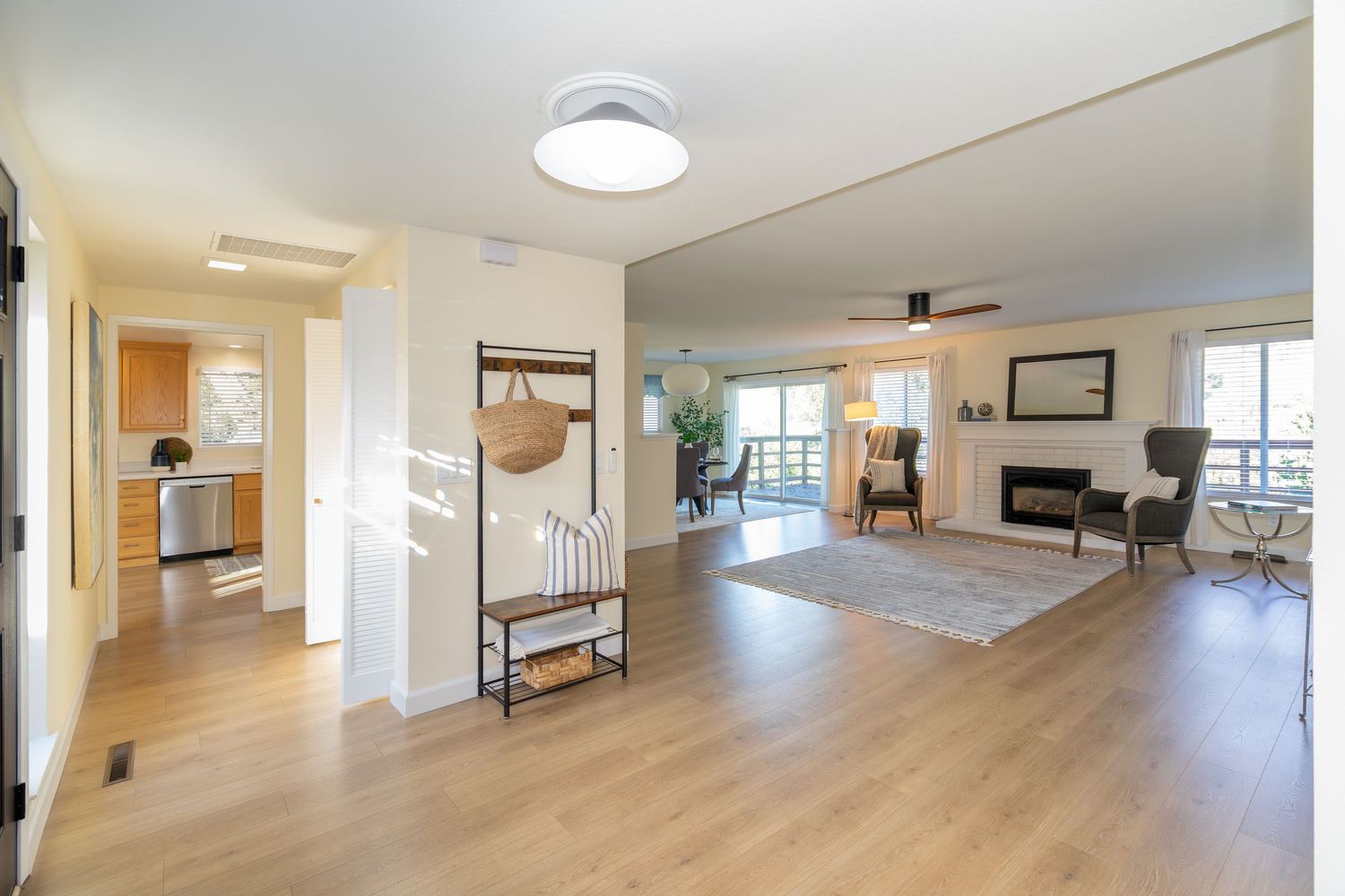 Bright open-plan living area with light hardwood floors, a fireplace, ceiling fan, and a hallway leading to a modern kitchen.