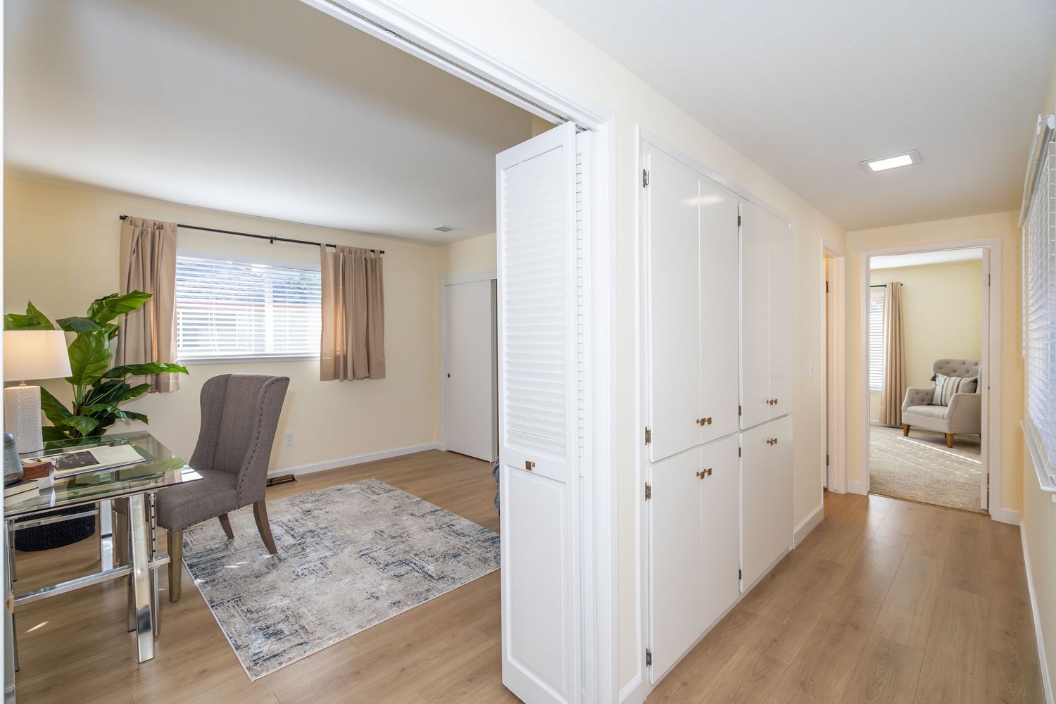 Bright hallway with white built-in cabinets, hardwood floors, leading to a home office with gray chair and rug.