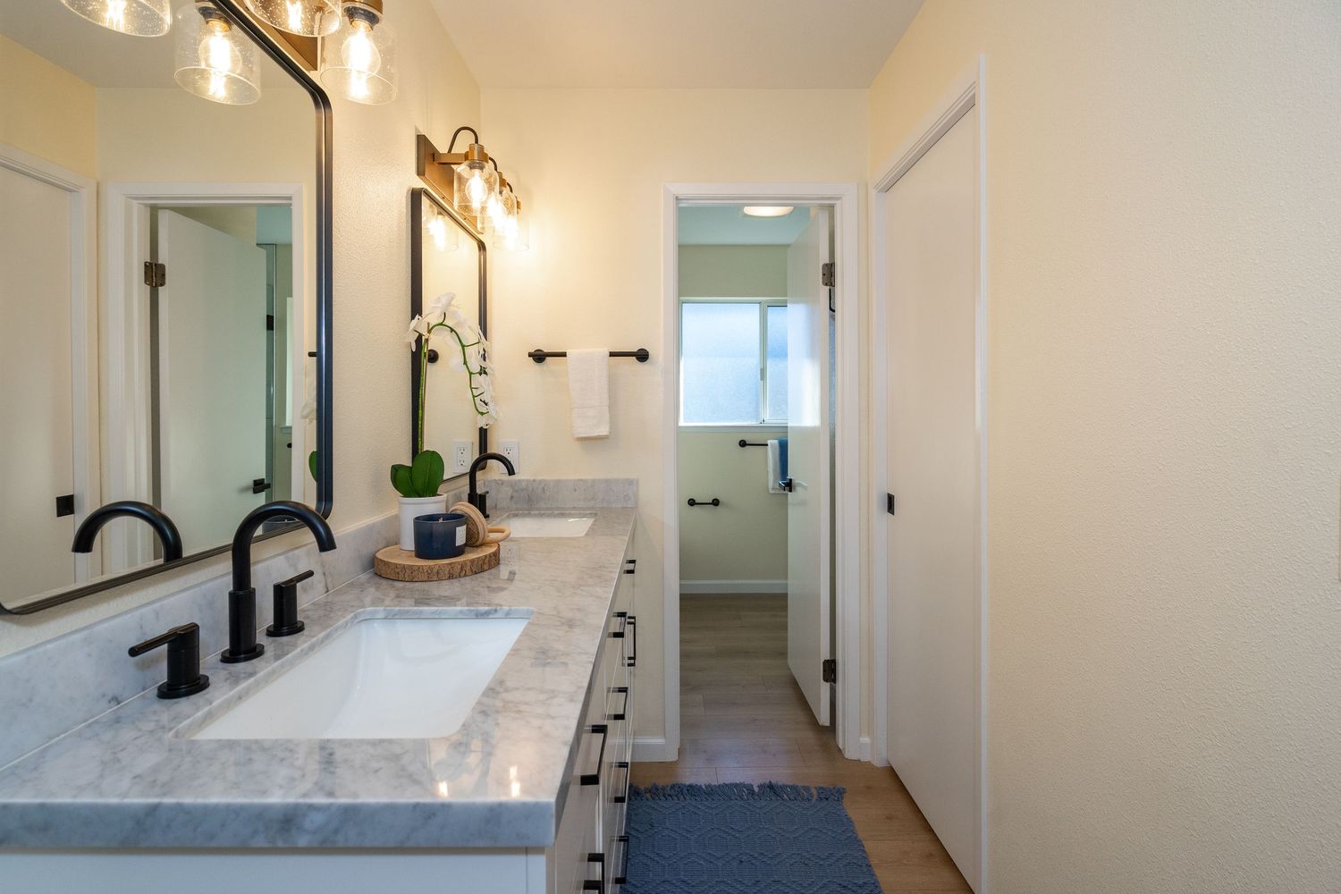 Modern bathroom with double marble vanity, black fixtures, large mirrors, and a hallway leading to a bright window.