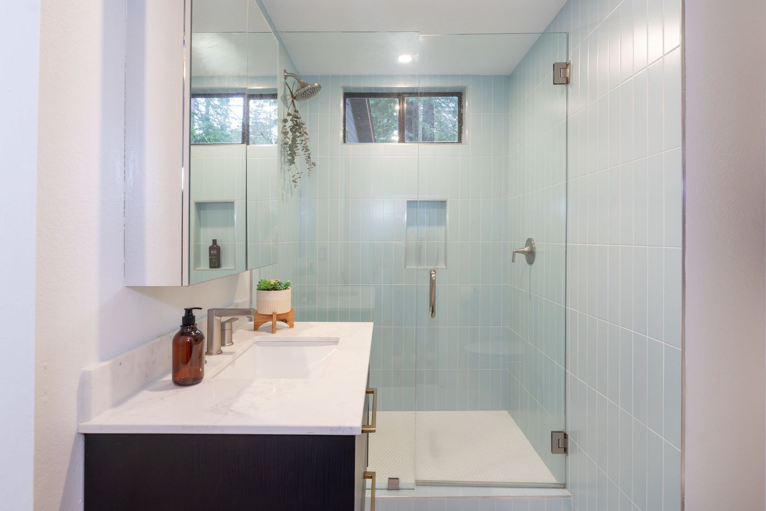 Modern bathroom with glass walk-in shower, light blue tiles, dark vanity, white countertop, and small window above.