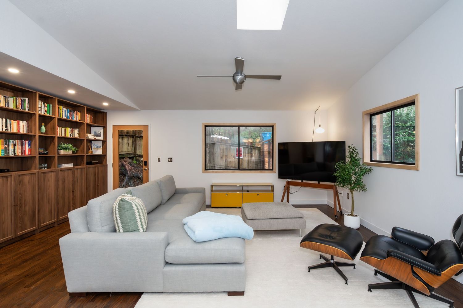 Modern living room with gray sectional sofa, Eames lounge chair, wall-mounted TV, built-in bookshelves, and skylight.