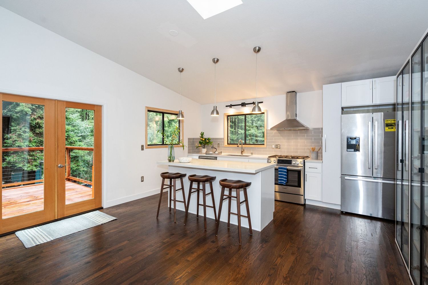 Modern kitchen with white cabinets, stainless steel appliances, island with bar stools, dark hardwood floors, and French doors to deck.