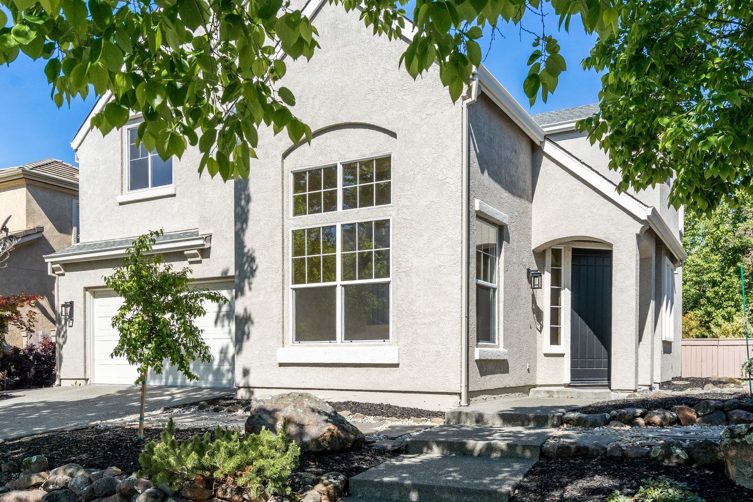 White stucco two-story home with arched window, dark front door, and lush green tree in foreground on sunny day.