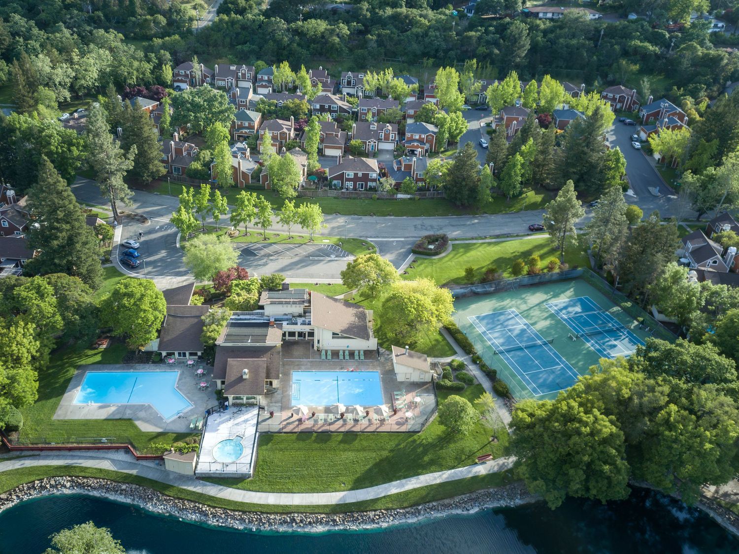 Aerial view of a luxury residential community featuring outdoor pools, tennis courts, and lush green trees beside a waterfront.