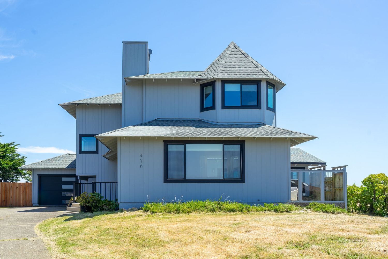 Gray two-story coastal home with blue windows, peaked roof, chimney, and manicured lawn under a bright blue sky.