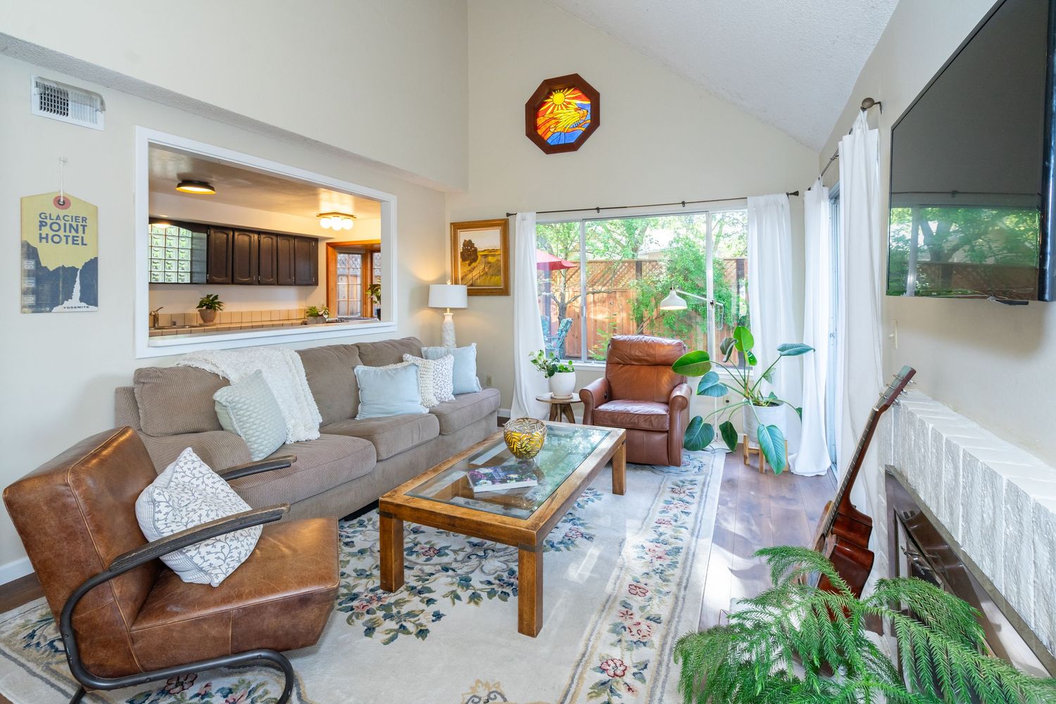 Bright living room with vaulted ceilings, leather sofa, glass coffee table, patterned rug, and large windows overlooking lush greenery.