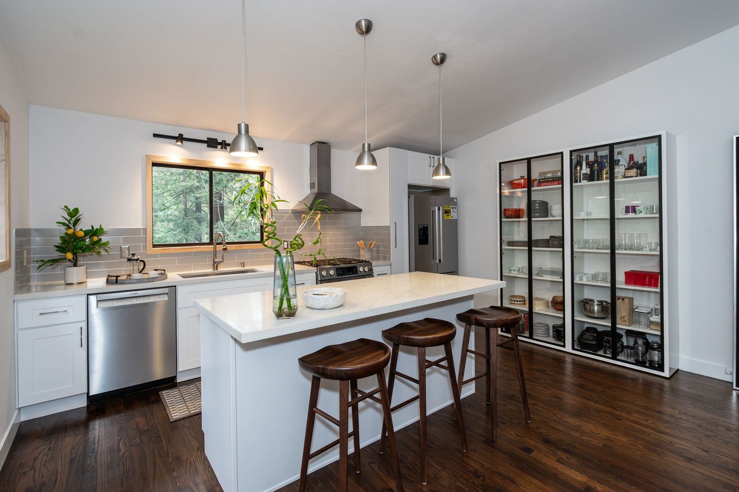 Modern white kitchen with island, bar stools, pendant lights, stainless appliances, and dark hardwood floors.