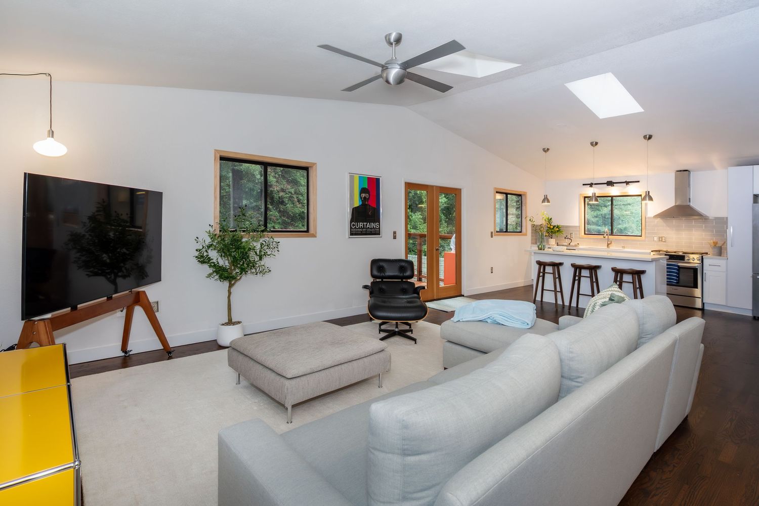 Modern open-concept living room with gray sectional sofa, ceiling fan, skylight, and kitchen visible in background.