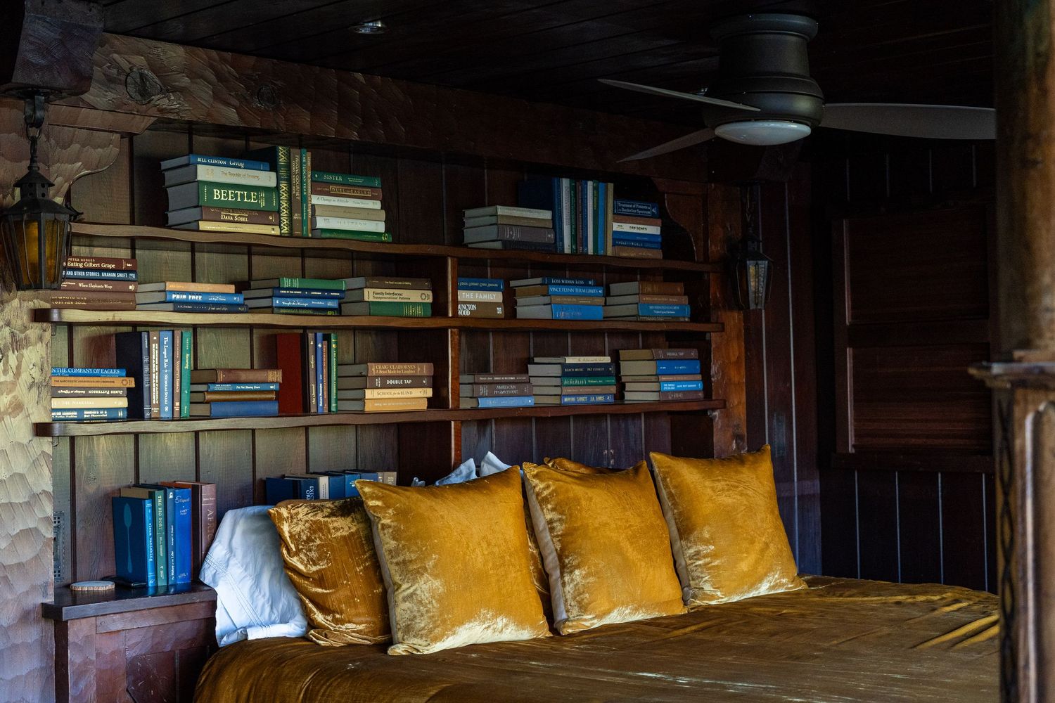 Cozy rustic bedroom with golden velvet pillows on a bed, wooden walls, and shelves filled with colorful books.