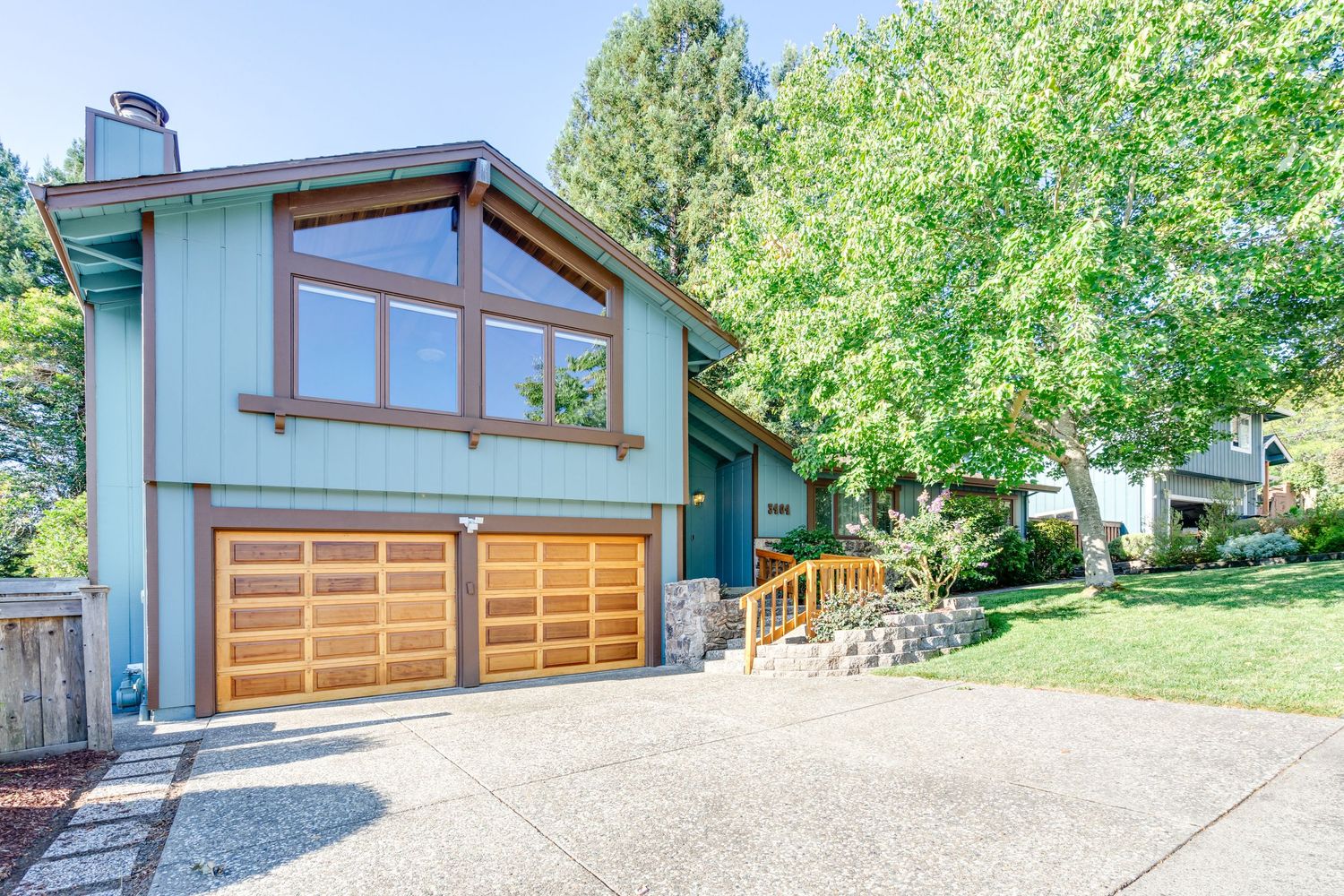 Blue two-story home with wood garage doors, large windows, and a landscaped driveway surrounded by mature trees.