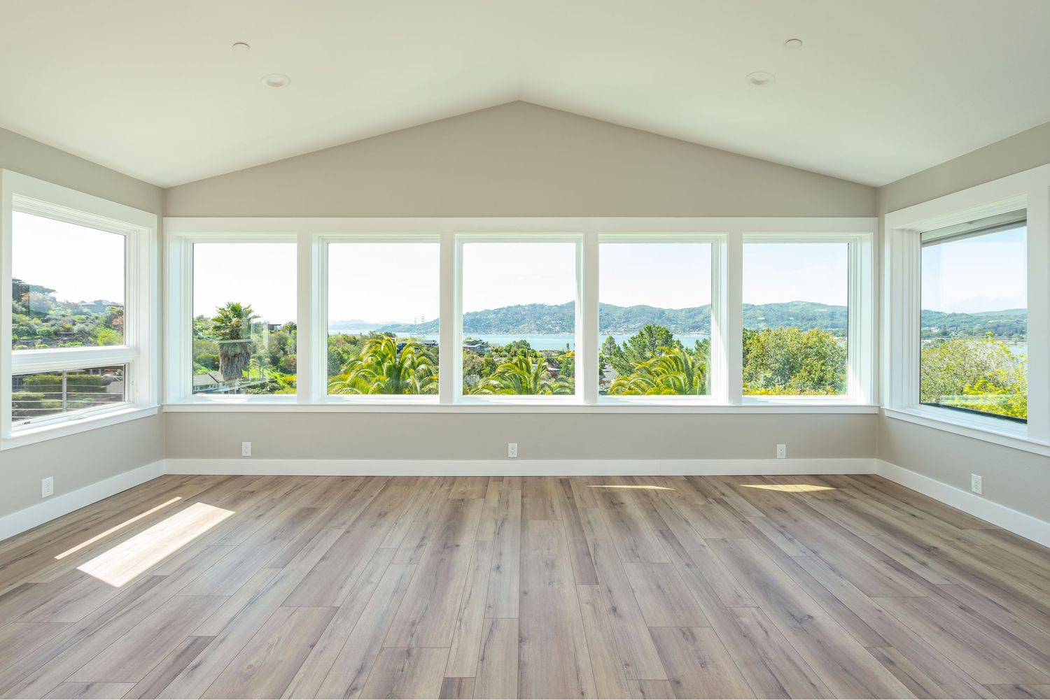 Bright empty room with vaulted ceiling, hardwood floors, and large windows overlooking lush green trees and scenic hills.