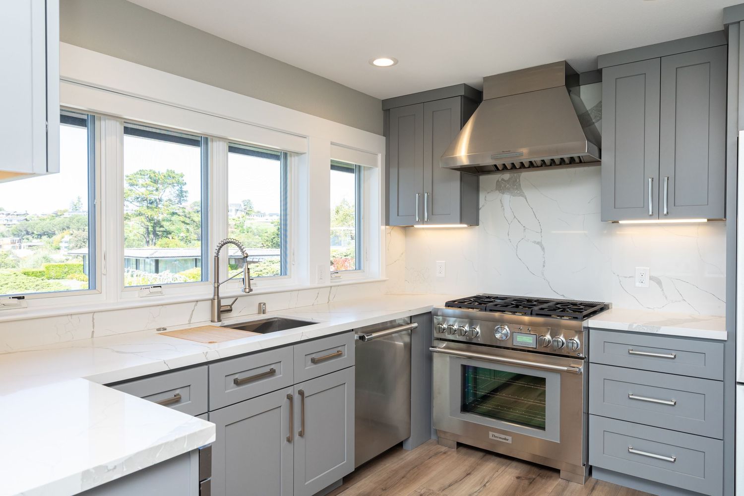 Modern kitchen with gray cabinets, stainless steel appliances, marble backsplash, and hardwood floors near bright windows.
