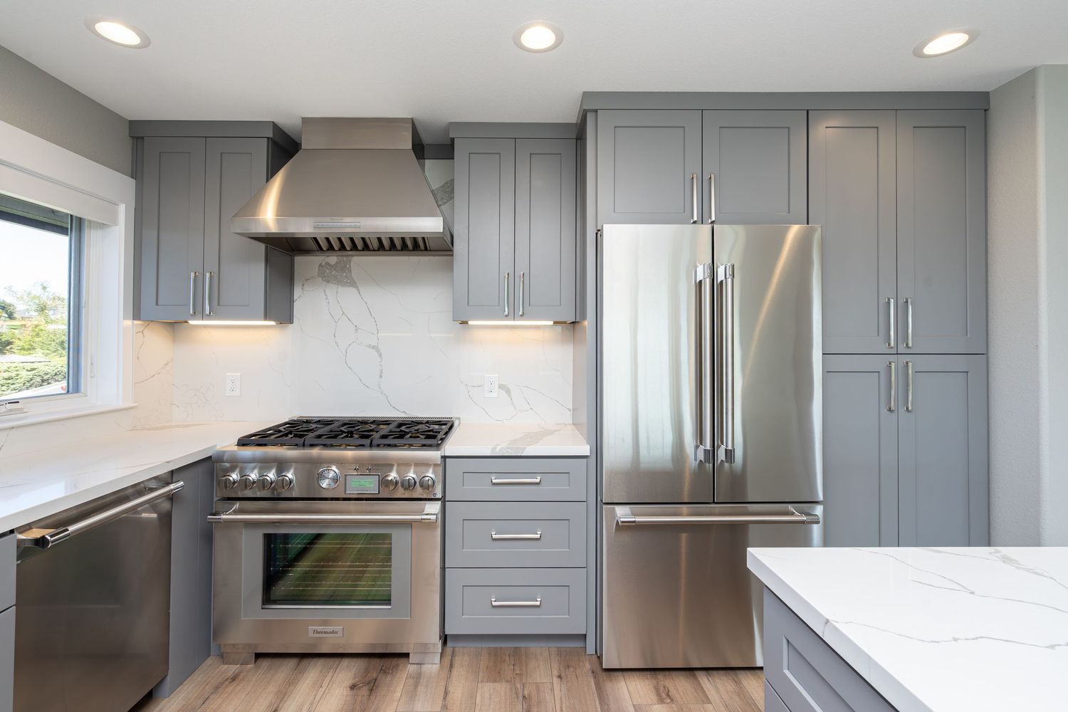 Modern kitchen with gray shaker cabinets, stainless steel appliances, marble backsplash, and hardwood floors.