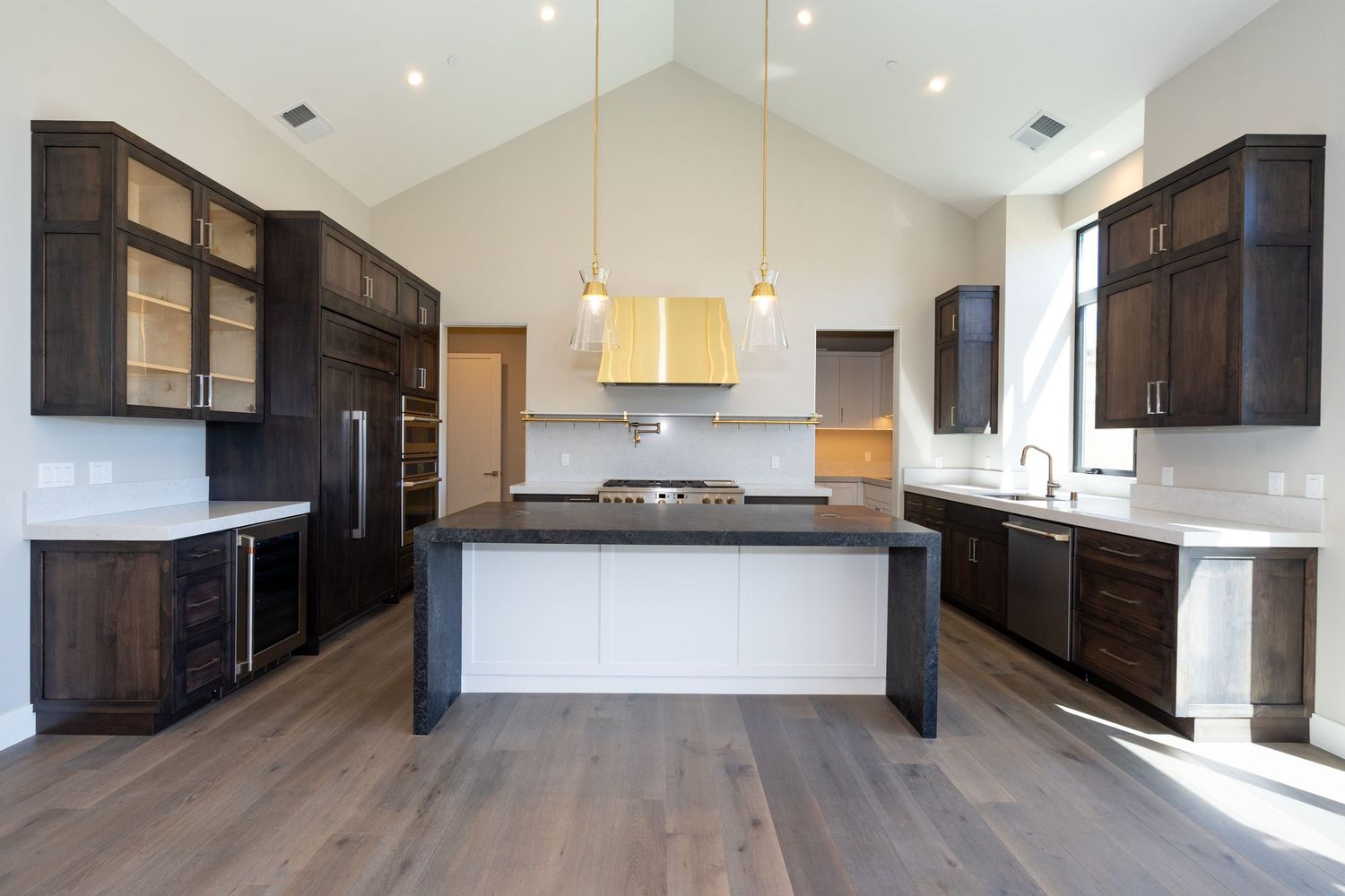 Modern kitchen with dark wood cabinets, large island, hardwood floors, and gold pendant lights under vaulted ceiling.