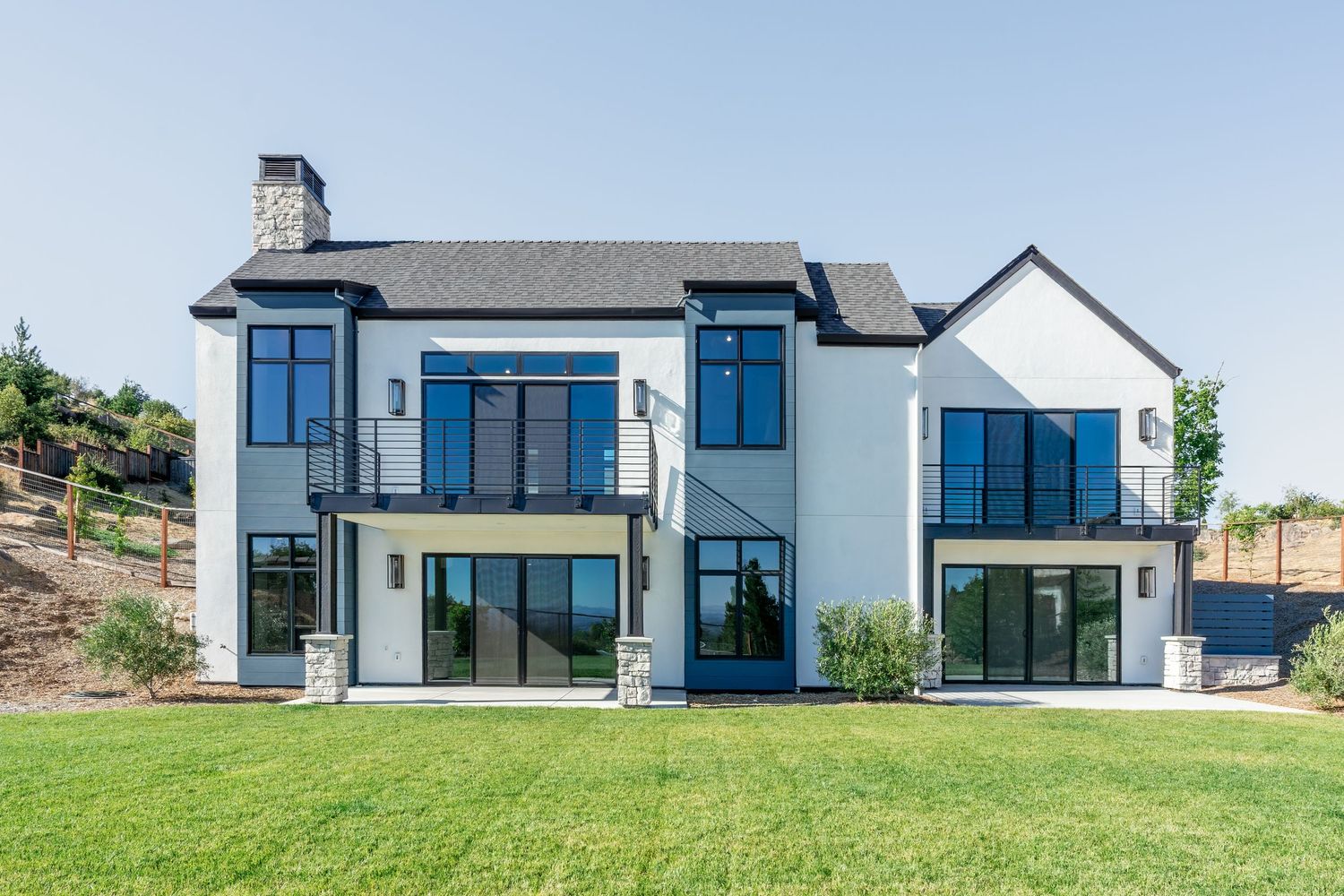 Modern two-story white home with black-framed windows, balcony, and lush green lawn on a sunny day.
