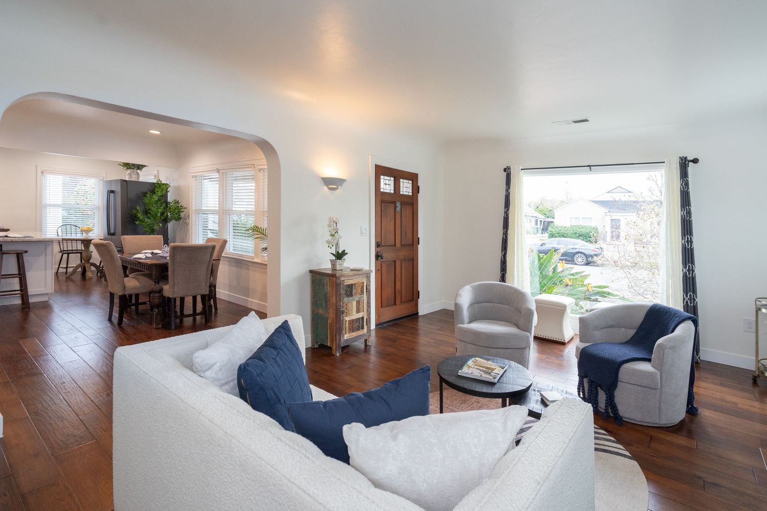 Bright living room with white sofa, hardwood floors, arched doorway leading to dining area, and large window with natural light.