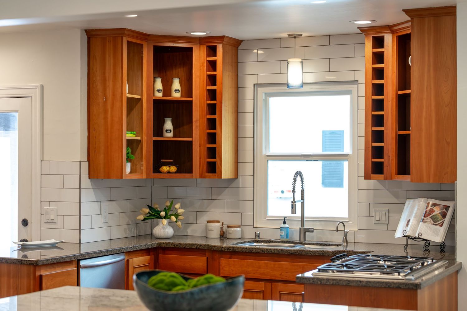 Modern kitchen with cherry wood cabinets, subway tile backsplash, stainless steel sink, and open shelving near window.