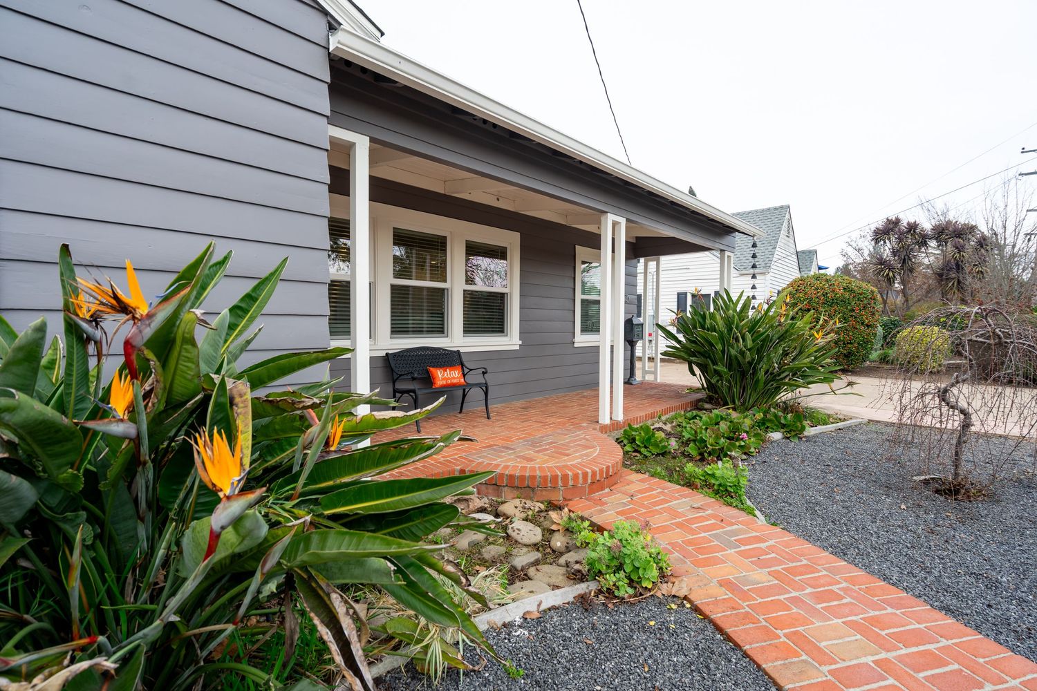 Gray house exterior with brick pathway, bird of paradise plants, and covered porch surrounded by lush landscaping.