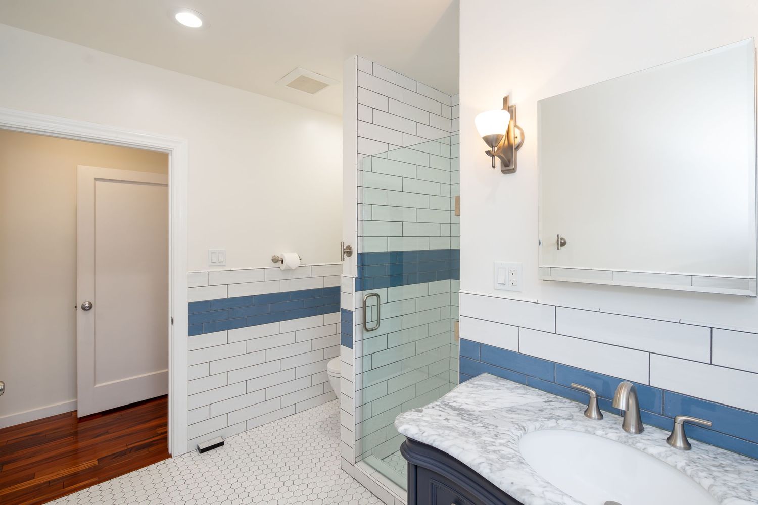 Modern bathroom with white subway tiles, blue accent stripe, marble vanity, walk-in shower, and hardwood floors.