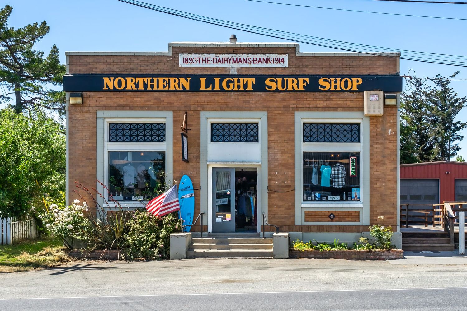Northern Light Surf Shop brick storefront with blue surfboard display and American flag on a sunny day.