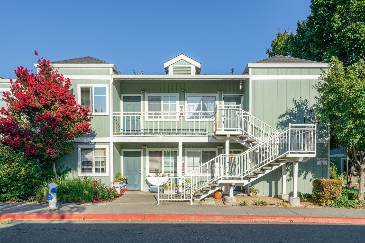 Two-story green townhouse with white balcony railings, exterior staircase, and red flowering tree in a sunny suburban setting.
