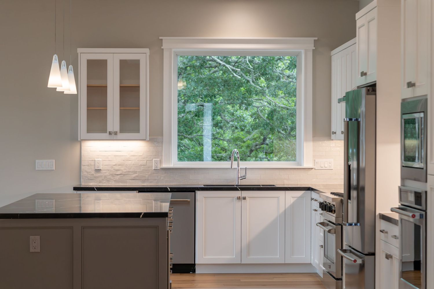 Modern white kitchen with black countertops, stainless steel appliances, glass-front cabinets, and a large window above the sink.