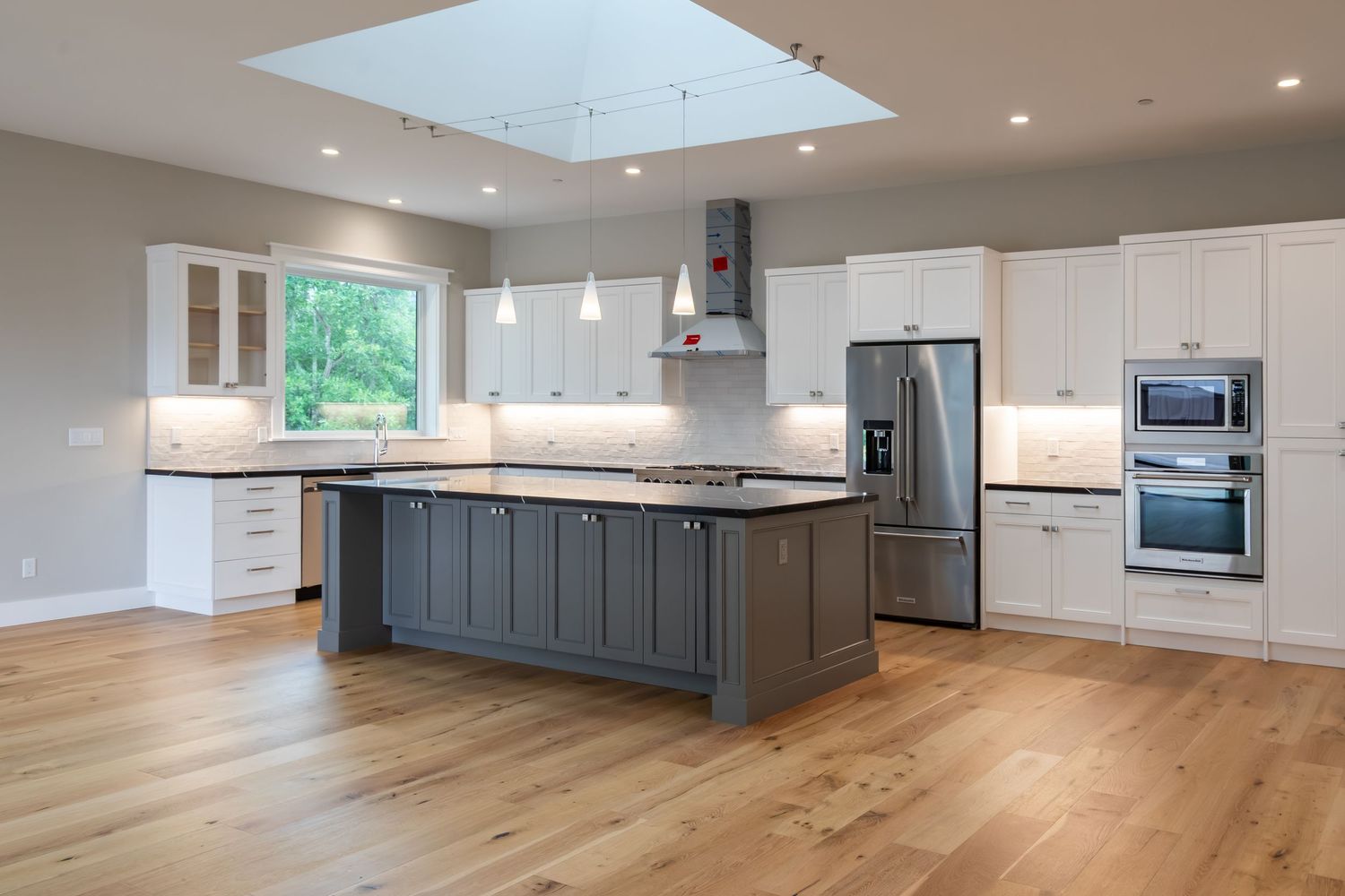 Modern kitchen with gray island, white cabinets, stainless steel appliances, and hardwood floors under a skylight.