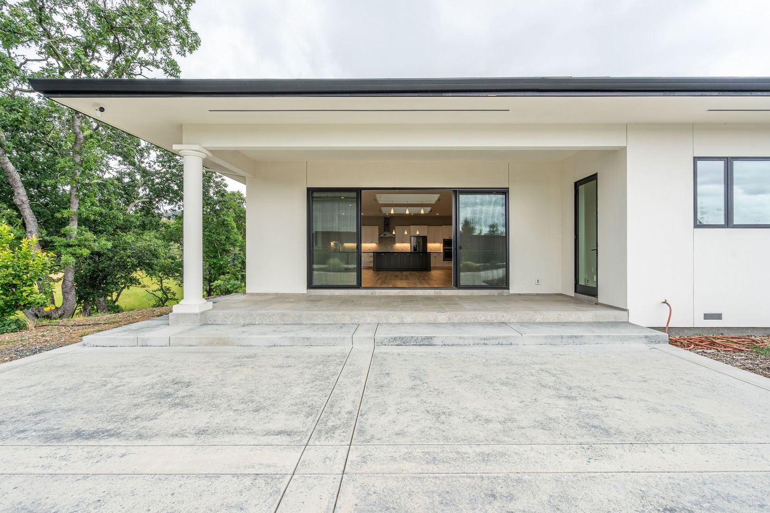 Modern white home exterior with large glass doors, concrete patio, and lush green trees in the background.