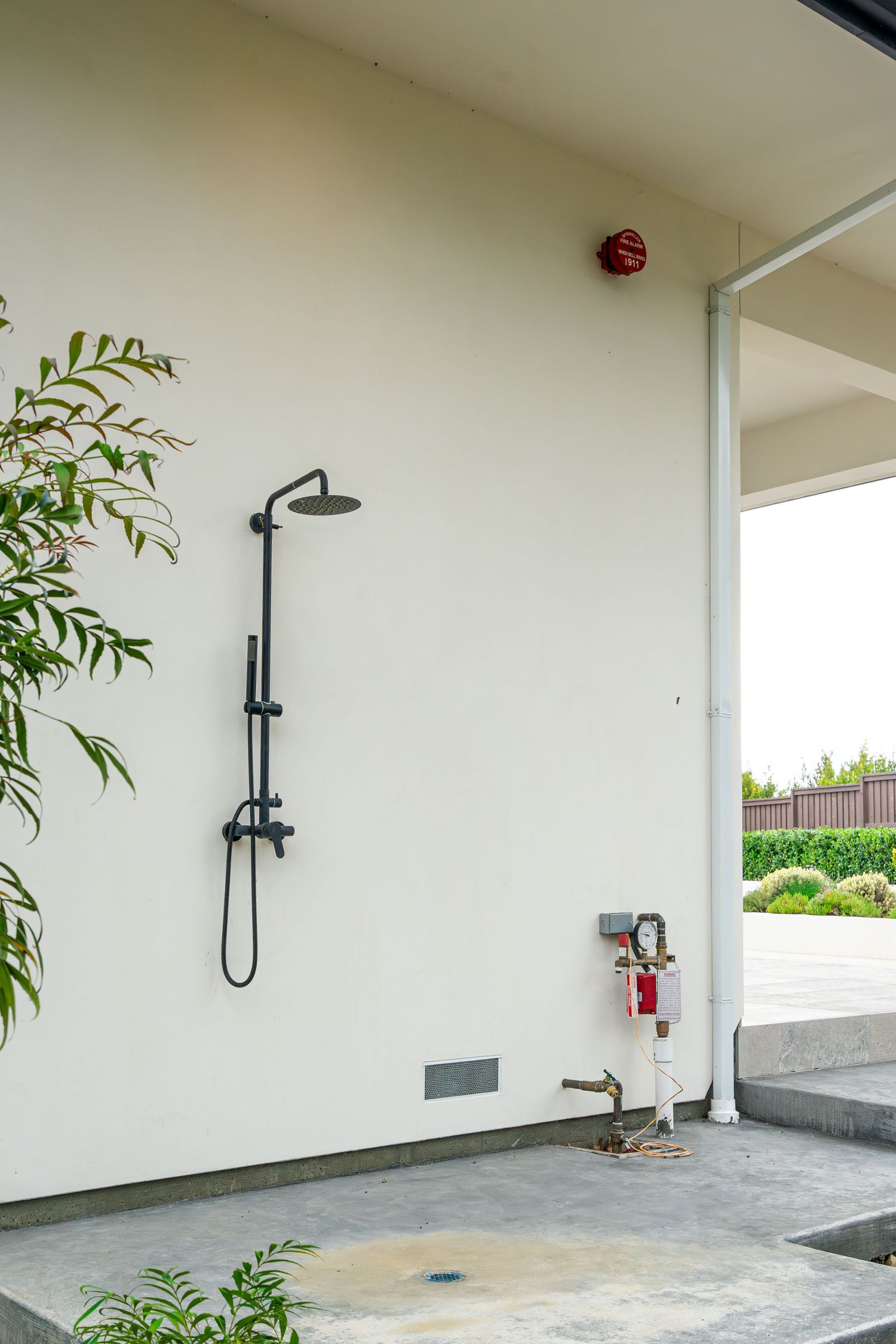 Black outdoor shower fixture mounted on white wall with hand shower, beside garden tap and lush green plants.