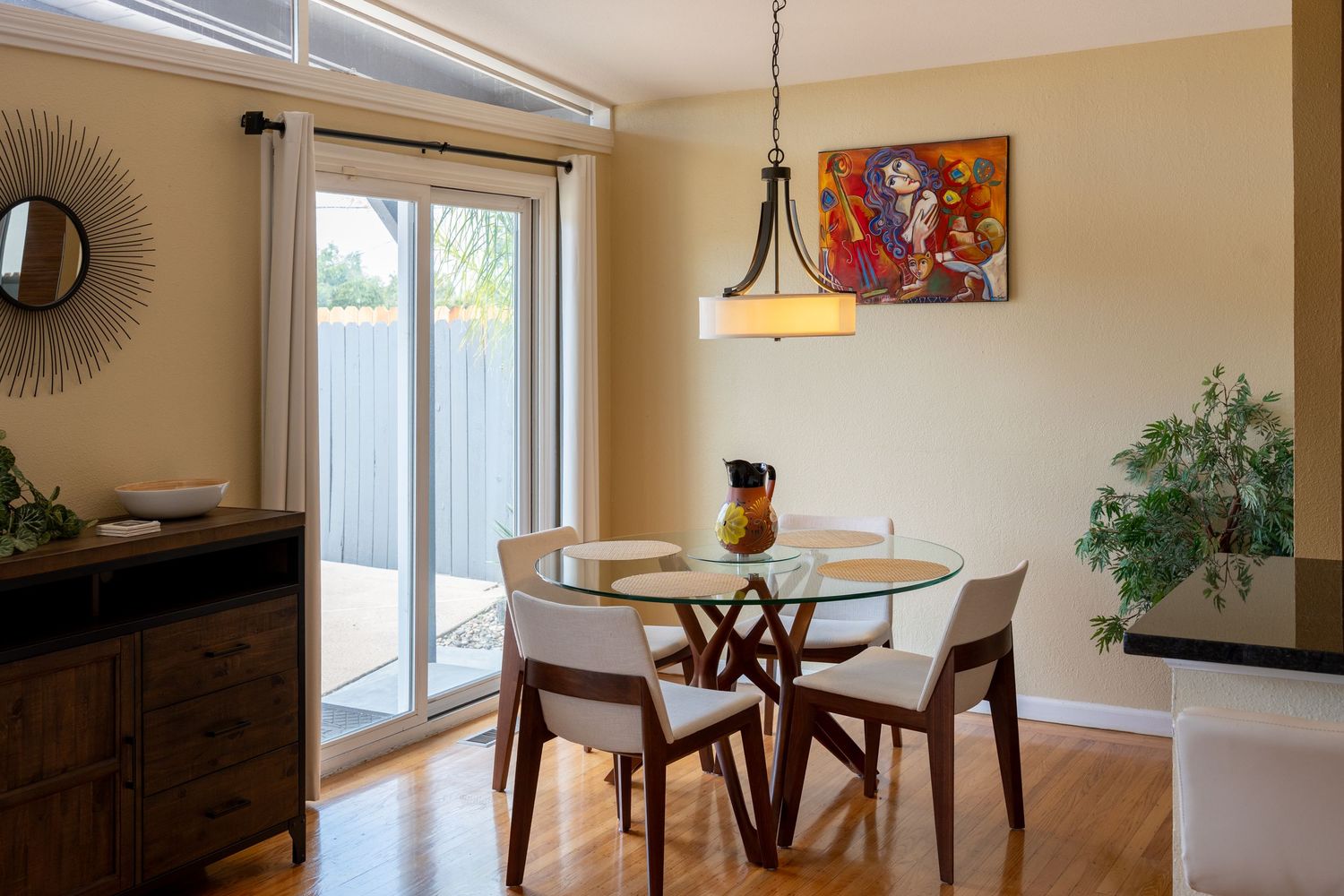 Bright dining room with glass round table, white chairs, pendant light, sliding glass door, and colorful wall art.