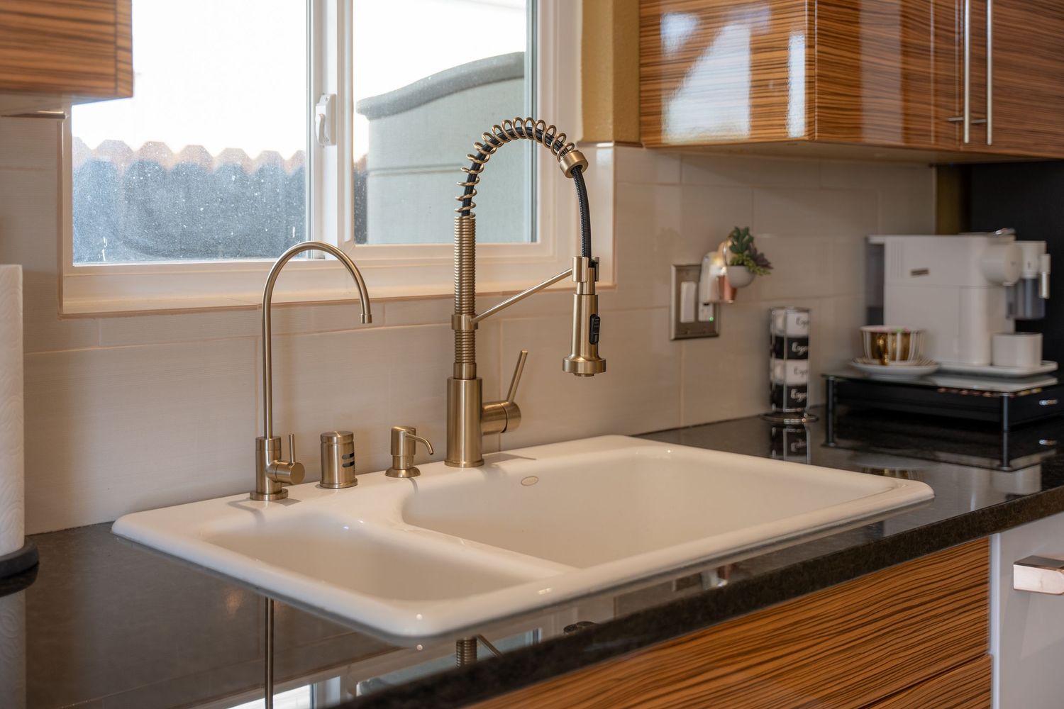 Modern kitchen sink with dual basins, brass faucet, and spring sprayer set in dark granite countertop near window.
