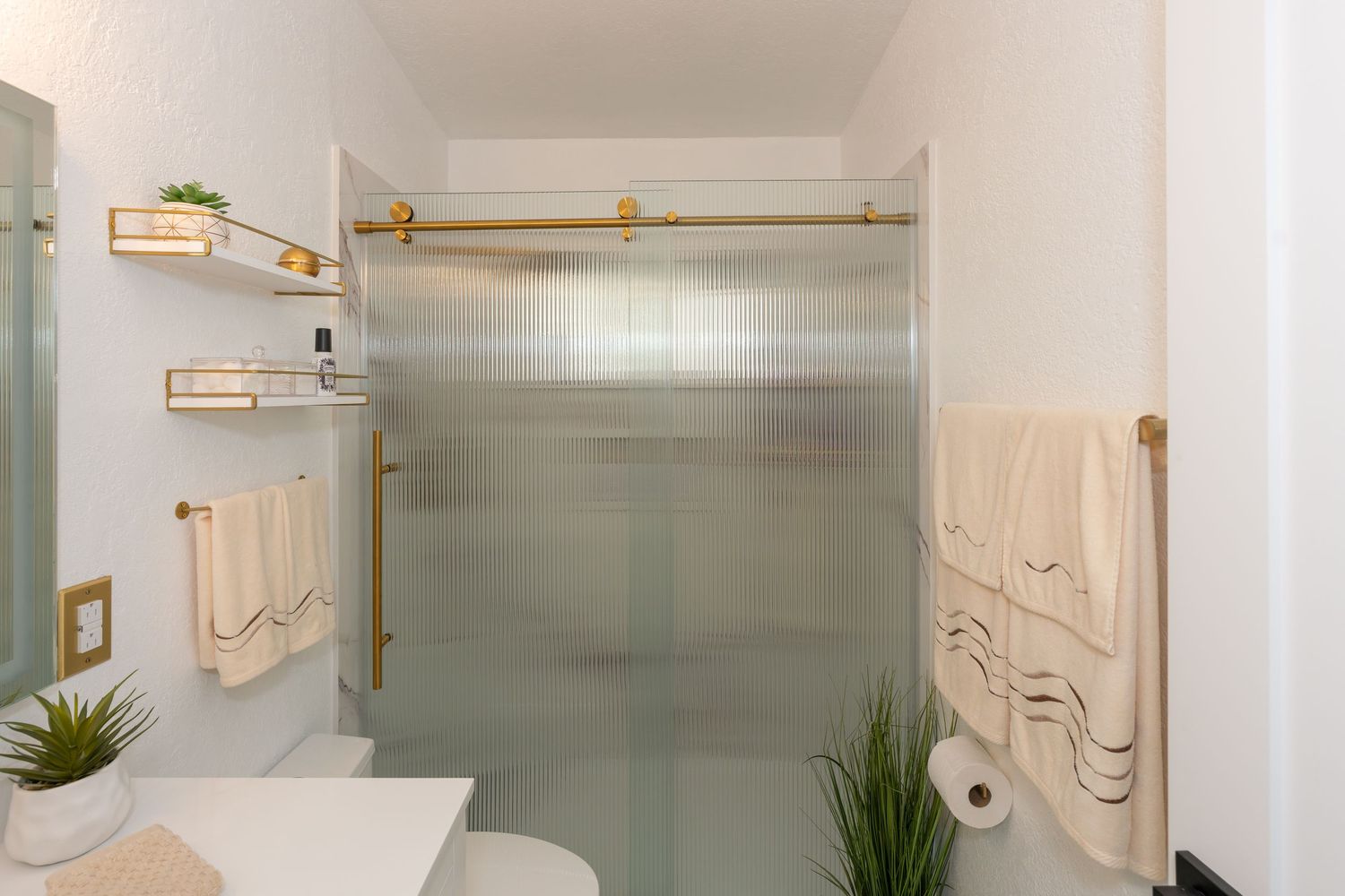 Modern bathroom with gold fixtures, frosted glass shower door, floating shelves, and neatly hung beige towels.