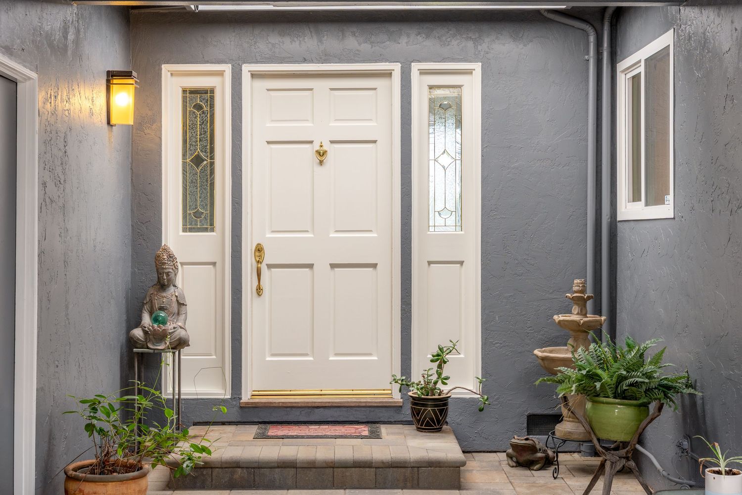 White front door with sidelights on a gray stucco home, flanked by potted plants and a decorative fountain.