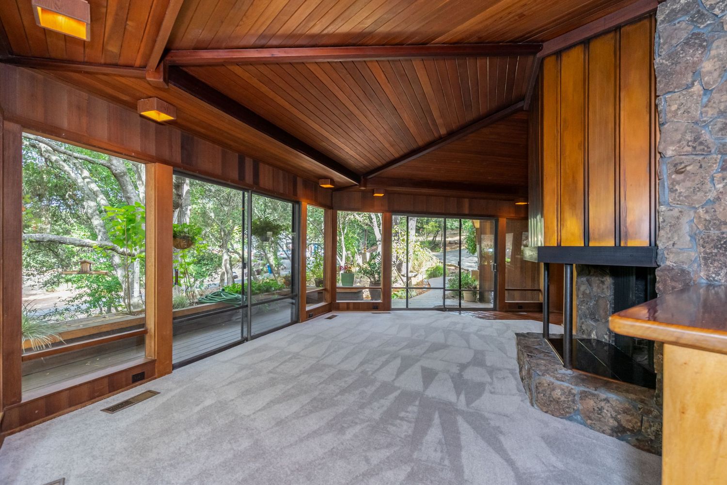Spacious mid-century living room with wood-paneled ceiling, floor-to-ceiling windows, stone fireplace, and lush garden views.
