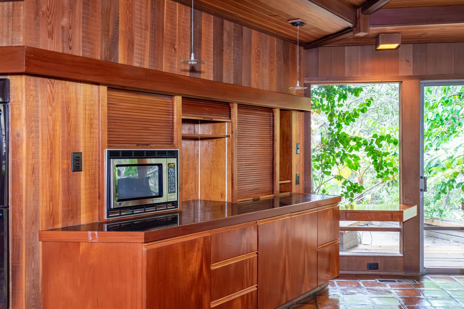 Warm wood-paneled kitchen with built-in microwave, rich mahogany cabinetry, granite countertop, and lush green garden view.