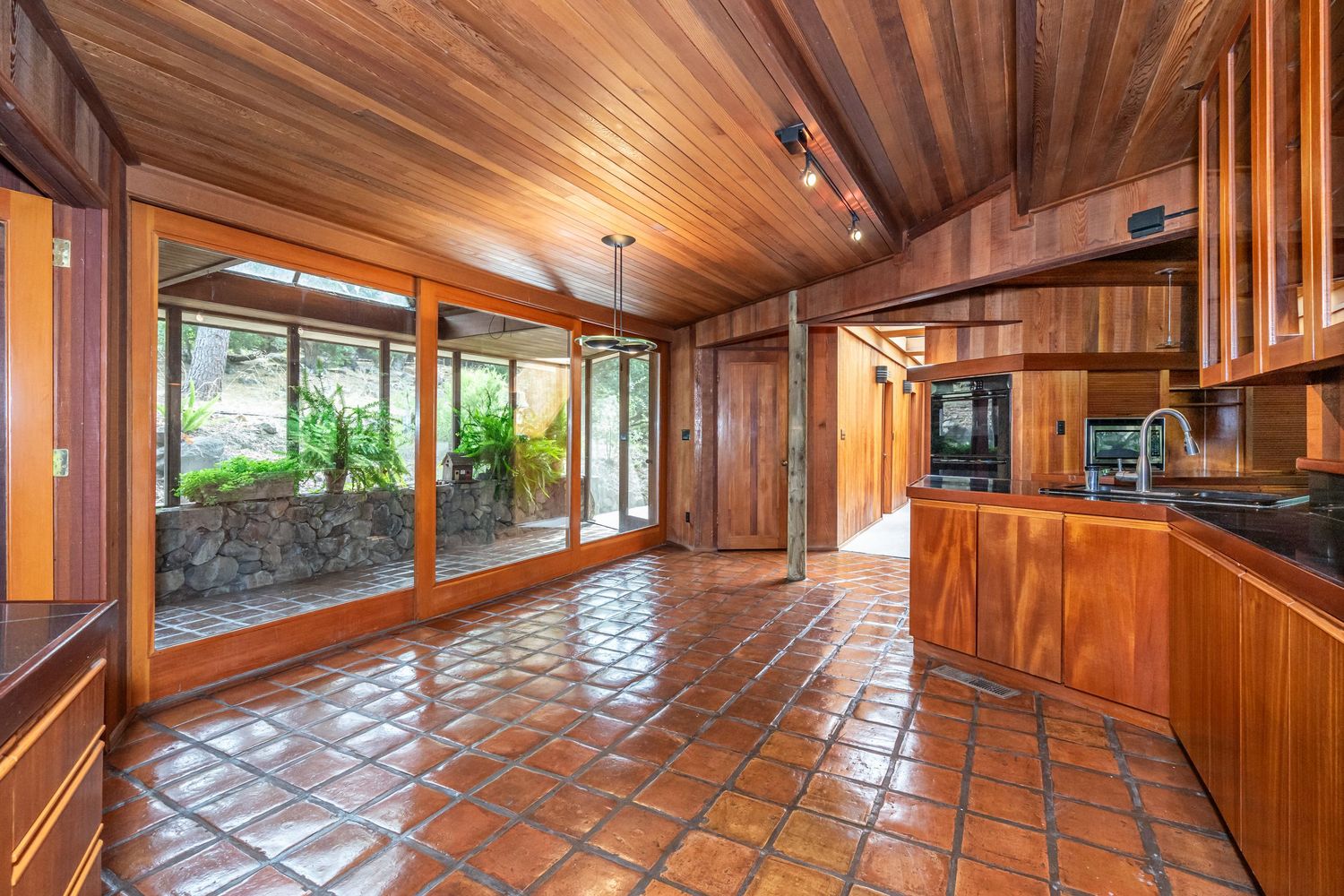 Spacious kitchen with rich wood cabinetry, terracotta tile floors, wood ceiling, and large sliding glass doors to lush greenery.