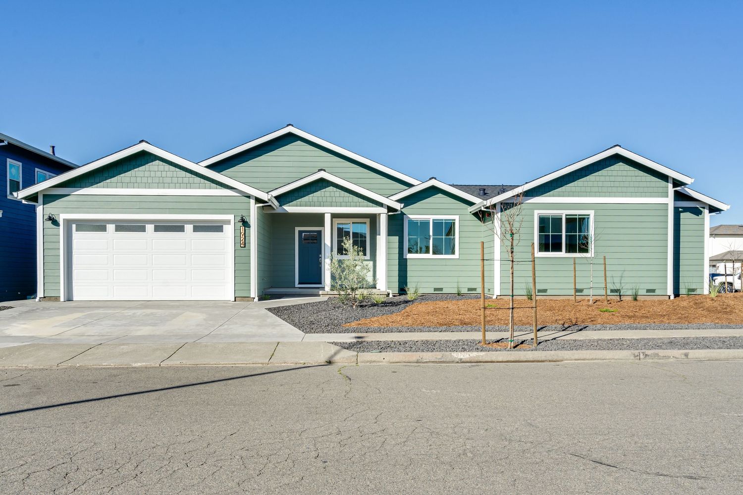 New construction single-story green home with white garage door, concrete driveway, and landscaped front yard under clear blue sky.