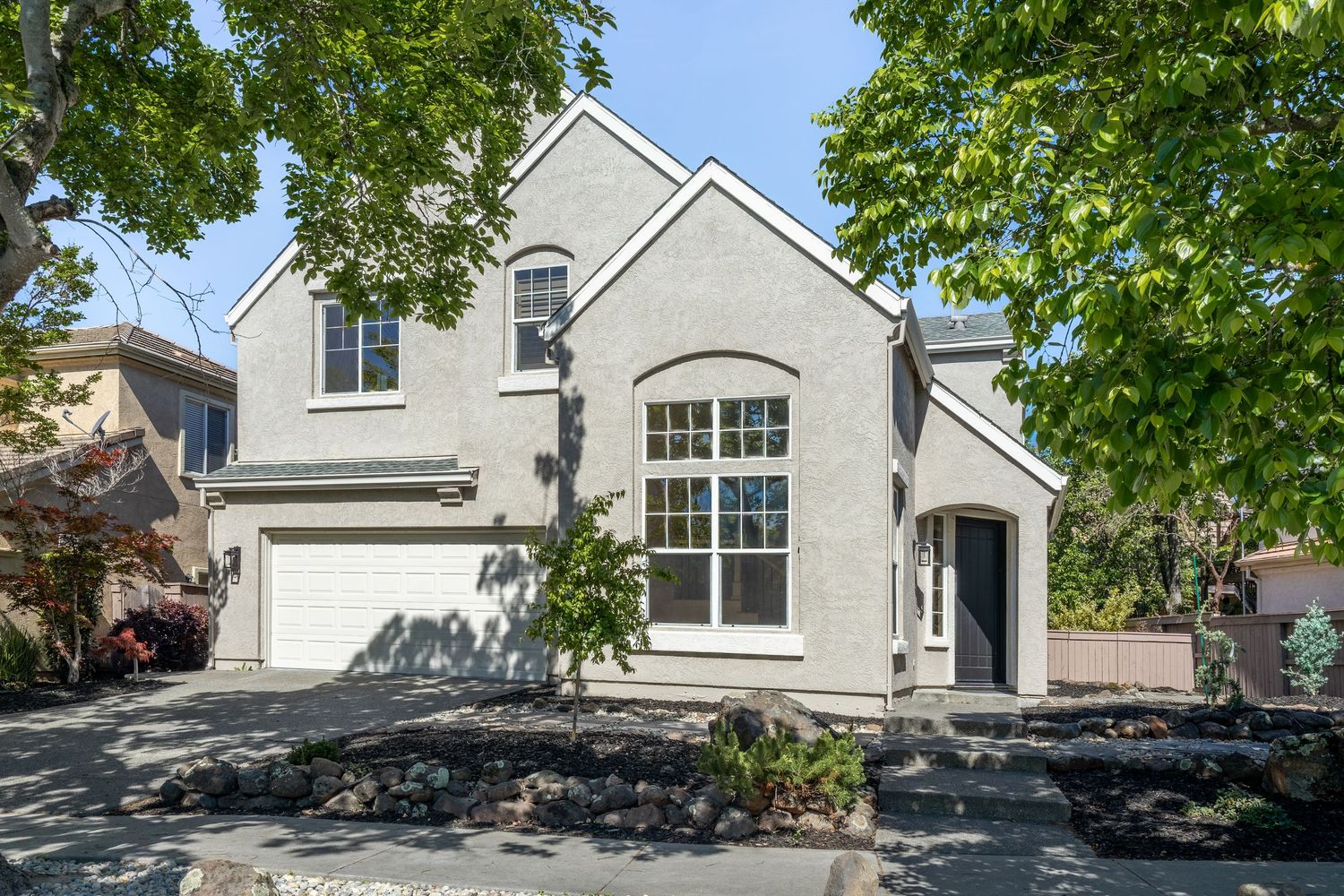 Two-story stucco home with arched window, attached garage, and mature trees in a suburban neighborhood.