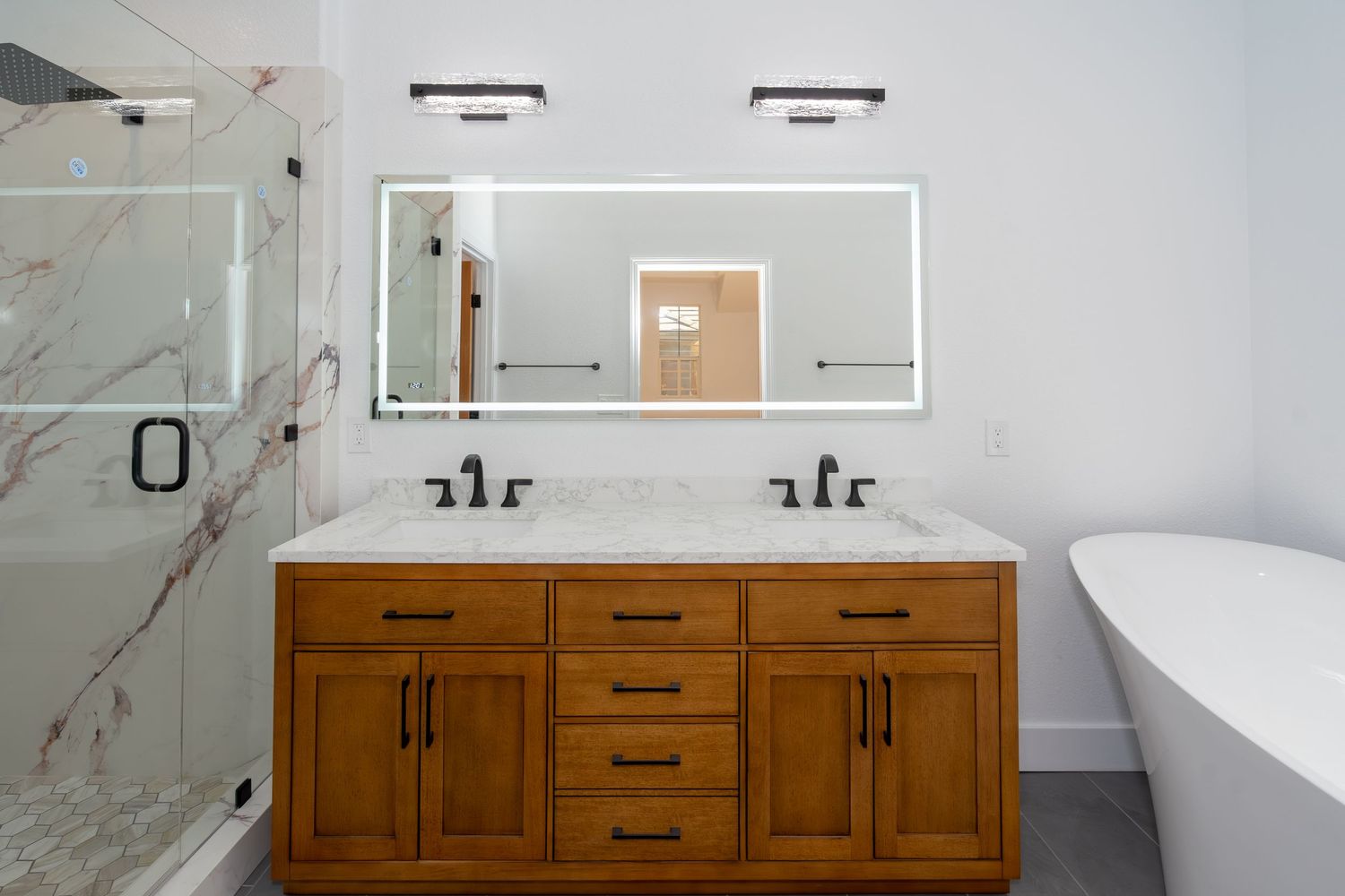 Modern bathroom with wooden double vanity, LED mirror, marble countertop, glass shower, and freestanding soaking tub.