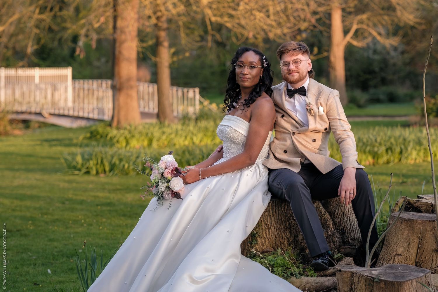 Interracial couple in wedding attire posing outdoors, bride in white gown holding bouquet, groom in gold tuxedo jacket.