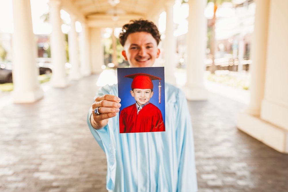 Graduate in blue cap and gown holds childhood kindergarten photo, smiling outdoors near white columns in redlands ca