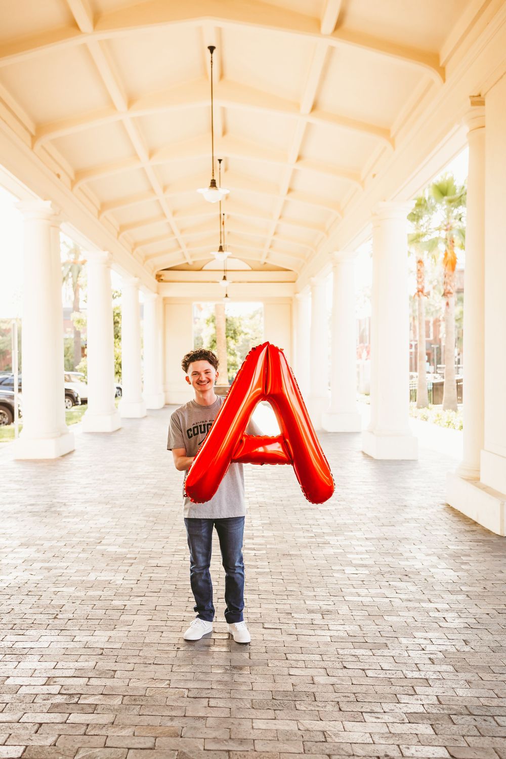 Smiling teen holds red letter A balloon in a white columned outdoor corridor with brick flooring.