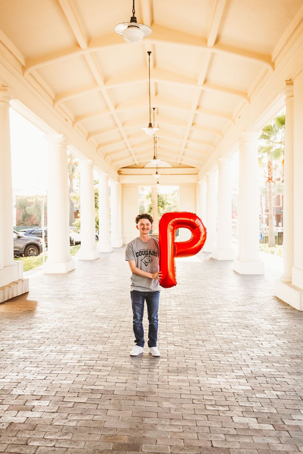 Young man holding a red letter P balloon in a elegant white columned corridor with brick flooring.