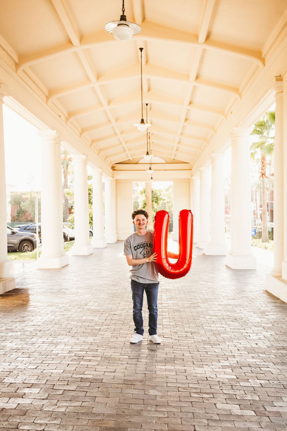 Young man smiling in a covered walkway holding a large red letter U balloon on brick pavers.
