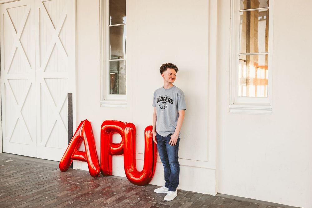Young man posing next to red APU letter balloons outside a white building on a brick pathway.