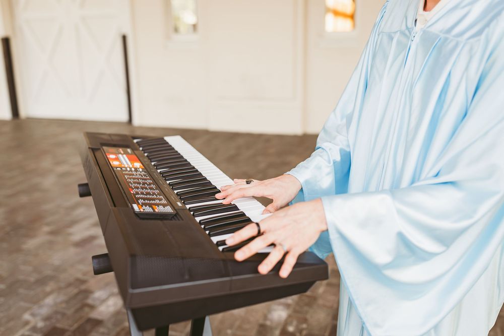 Person in light blue graduation gown playing a dark electronic keyboard piano indoors.