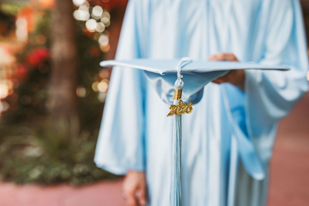 Graduate in light blue cap and gown holding diploma, with gold 2020 tassel, celebrating graduation day.