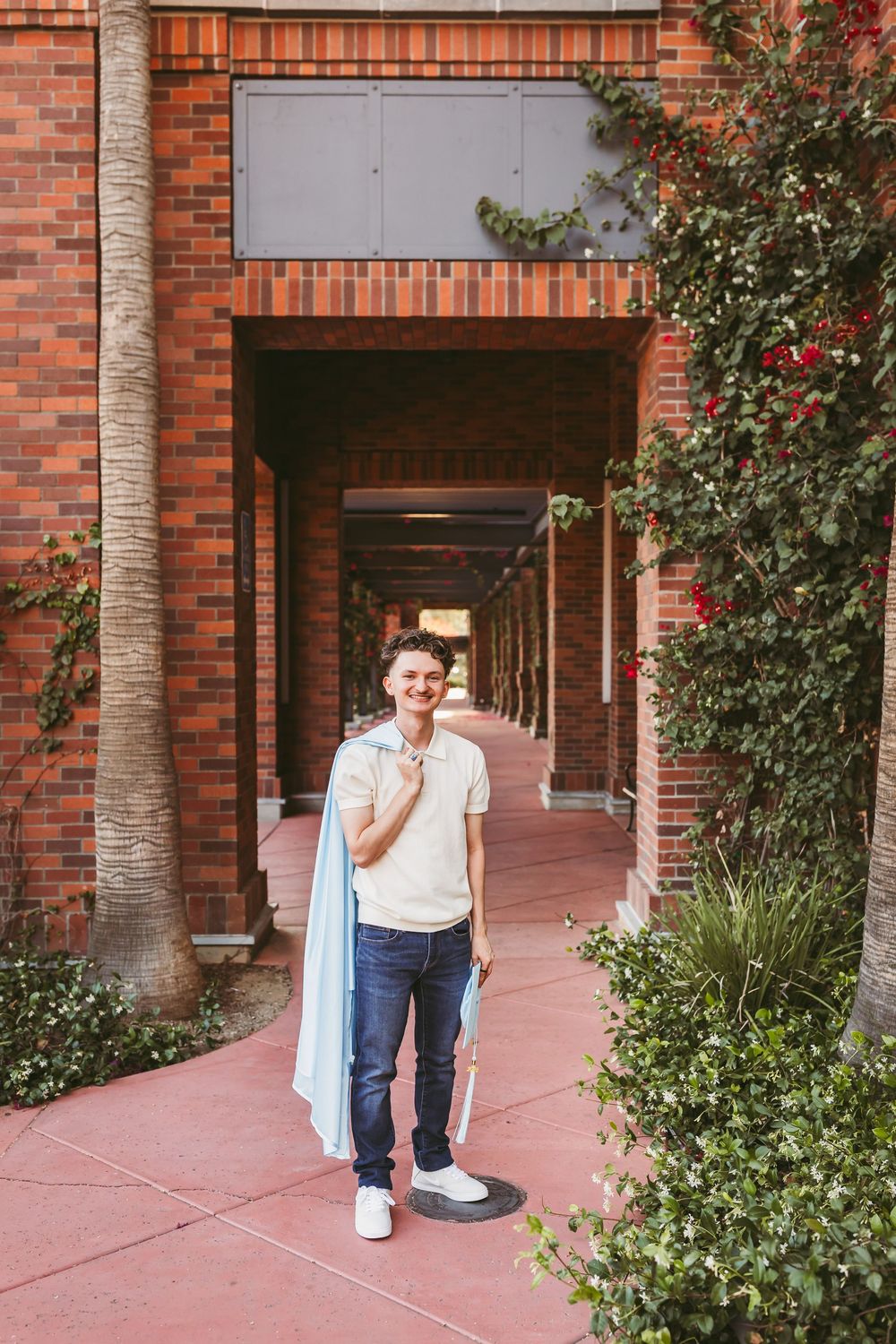 Young man smiling in a brick archway corridor with lush green ivy, wearing jeans and a white polo shirt.