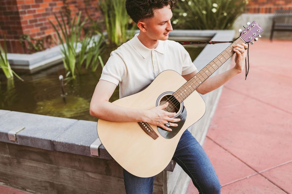 Young man playing acoustic guitar outdoors near a garden pond on a brick patio.